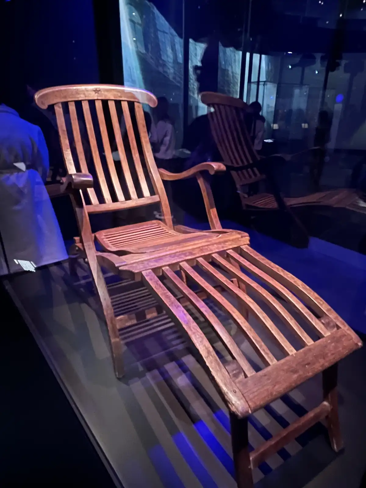a wooden lounge chair with a slatted back and footrest is displayed under blue lighting, likely in a museum or exhibit, with…