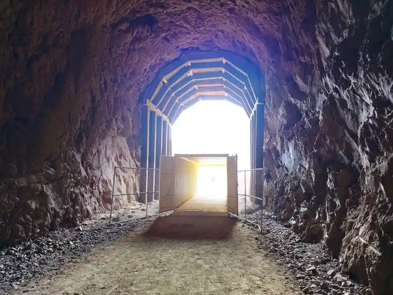 A view from inside a dark, rocky tunnel looking toward a bright, sunlit exit. The tunnel has a wooden arched roof structure…