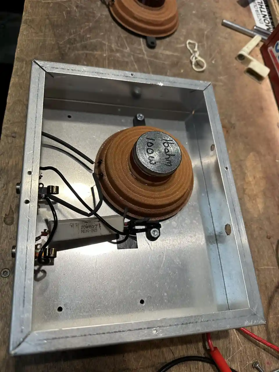 A close-up photo shows a homemade or modified speaker inside a metal enclosure on a workbench. The speaker cone has a…
