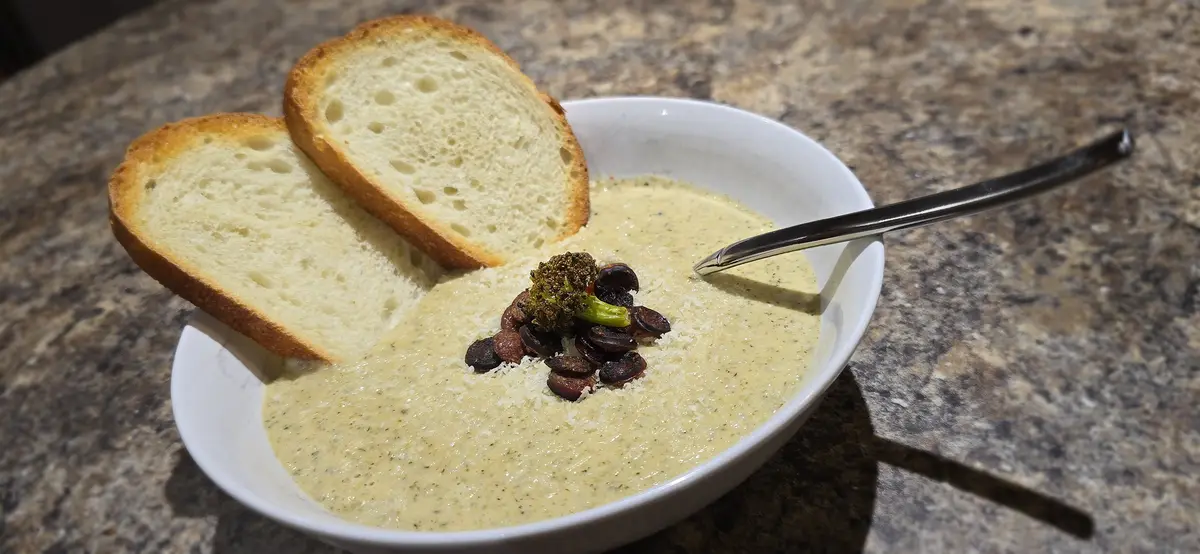 A white bowl of creamy soup sits on a speckled countertop, garnished with roasted broccoli, sliced mushrooms, and grated…