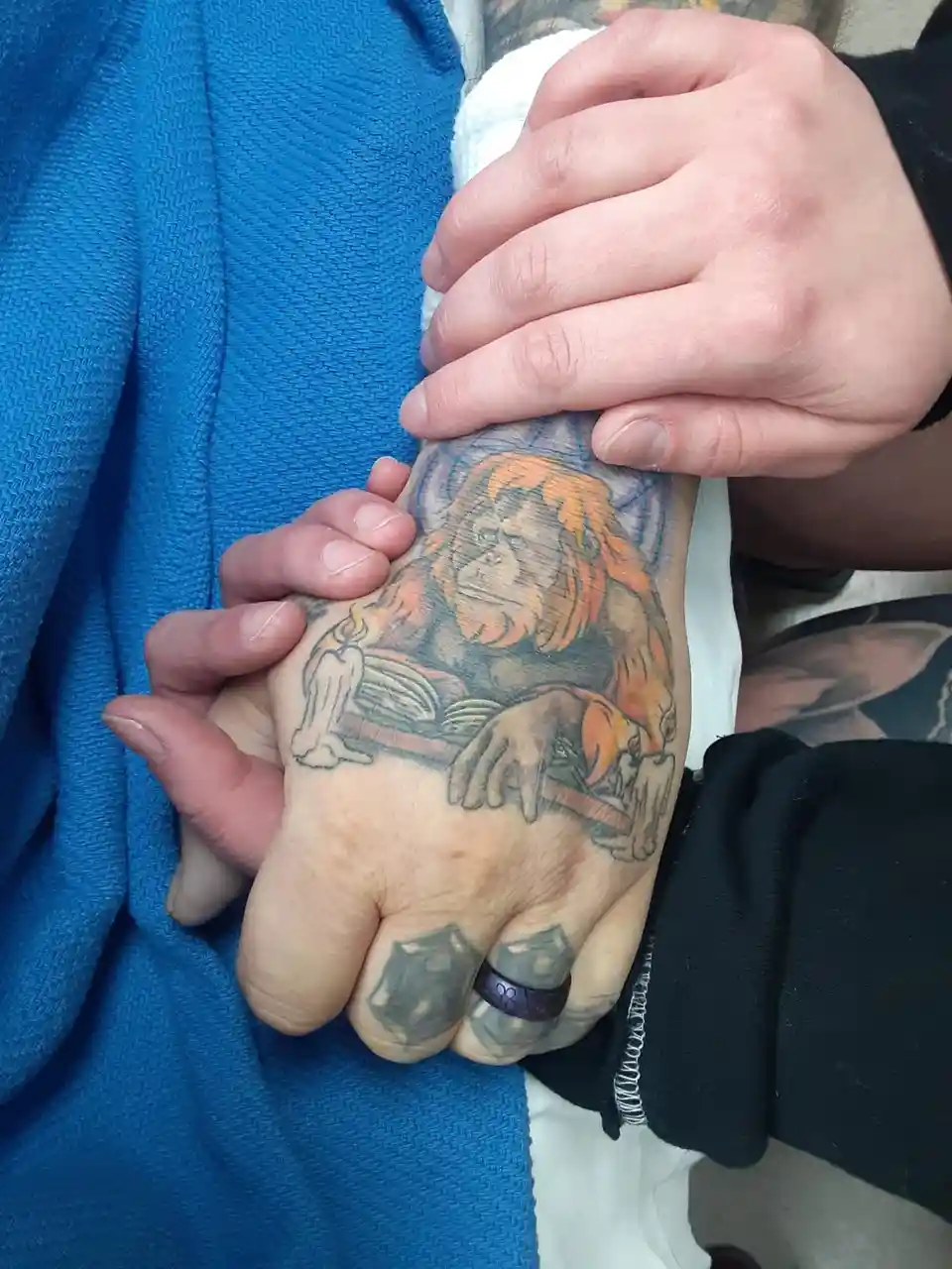 A close-up photo shows two hands clasped together, one hand displaying a colorful tattoo of a monkey with orange hair…