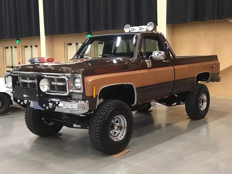 A brown and tan lifted classic Chevrolet pickup truck with oversized tires, chrome wheels, a custom front bumper, and…