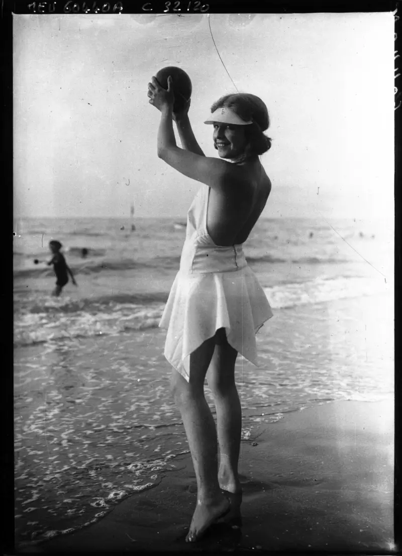 A vintage black-and-white photo shows a smiling woman in a white 1930s-style beach dress and visor standing in shallow ocean…