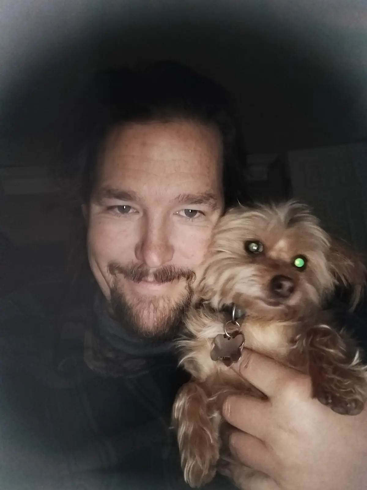 A man with a beard and dark hair smiles while holding a small, fluffy brown dog in a dimly lit room. The dog's eyes glow…