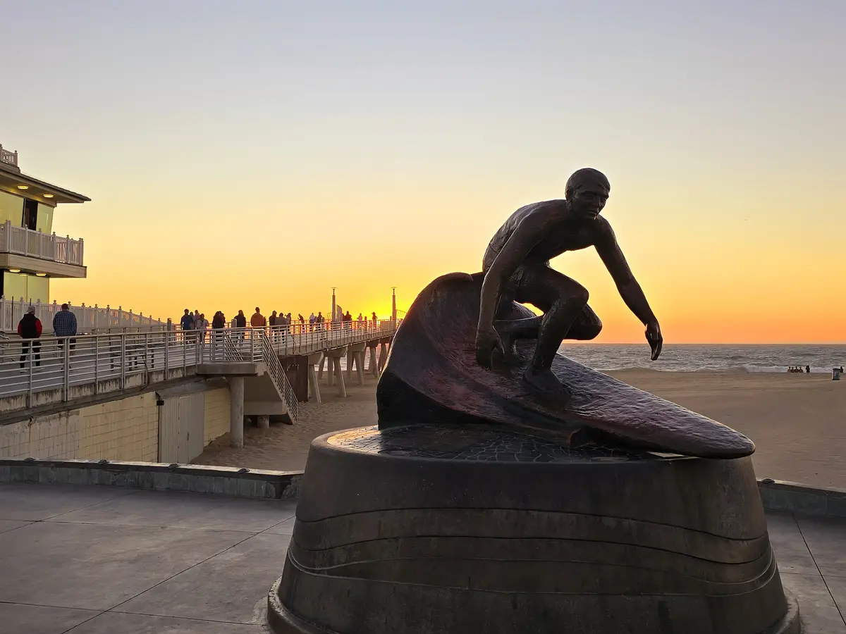 A bronze statue of a surfer riding a wave is prominently featured at sunset, with a pier and beach in the background. The…