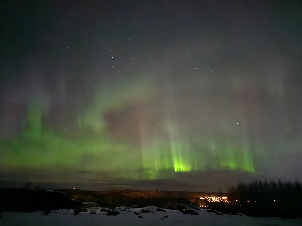 A long-exposure photograph captures vibrant green auroras dancing across a starry night sky above a snowy landscape, with…