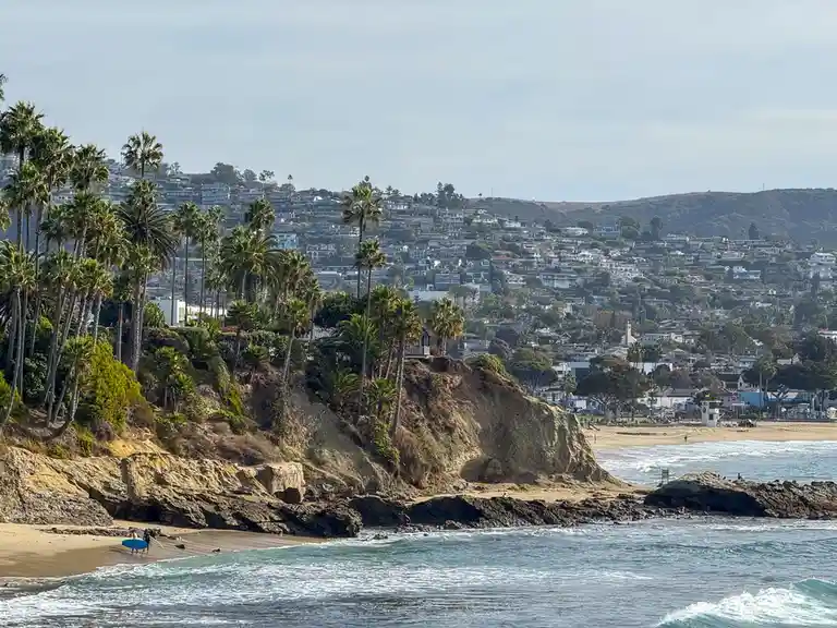 A scenic coastal view shows a beach with waves, cliffs, palm trees, and a hillside town in the background. Two people with…