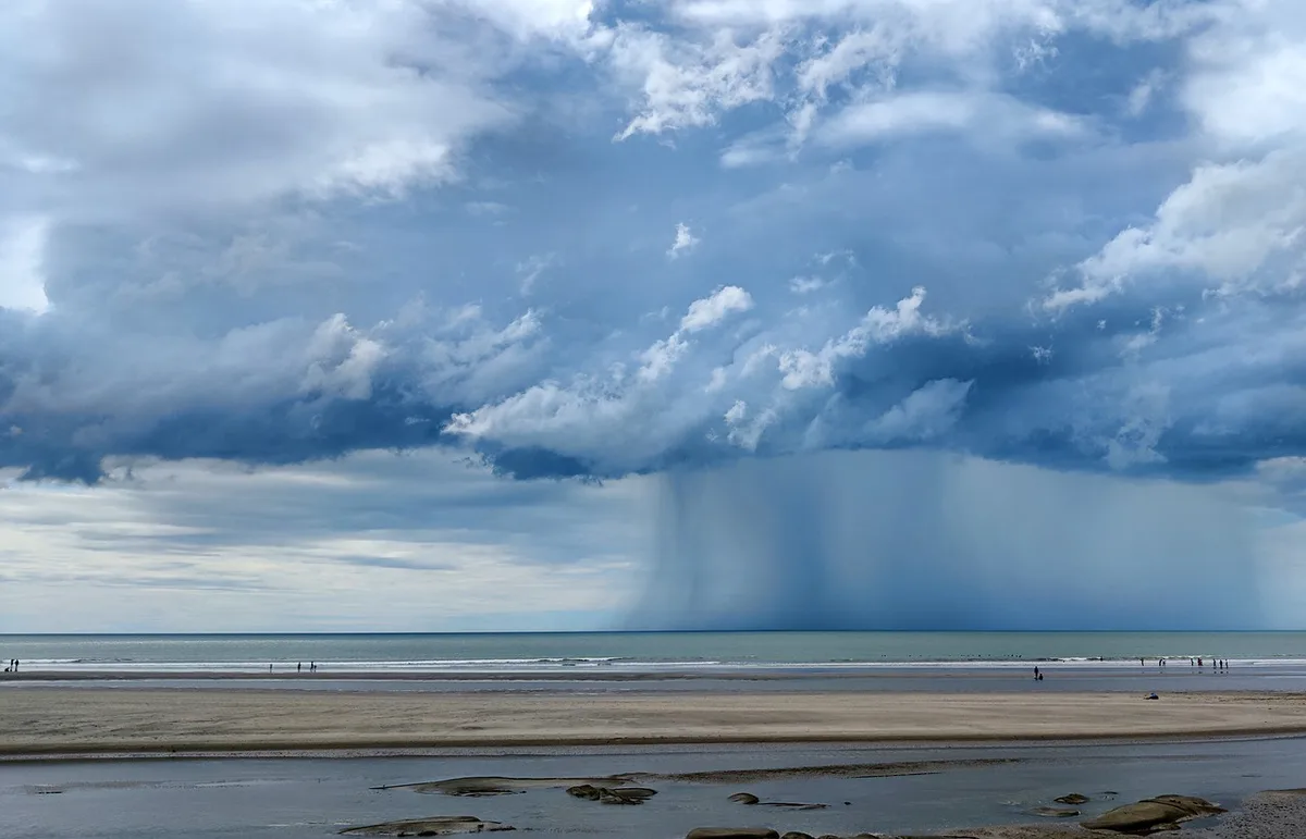 A wide beach scene with people scattered along the shoreline under a vast, dramatic sky filled with dark, heavy clouds. Rain…