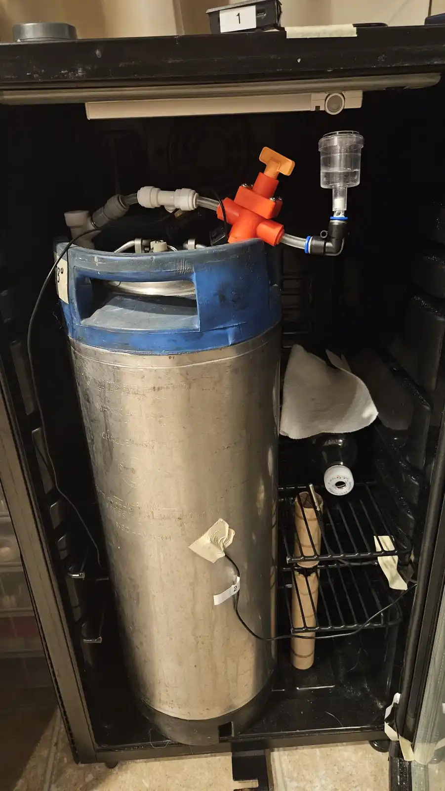 A large stainless steel keg with a blue plastic top sits inside a small black refrigerator, connected to orange and clear…