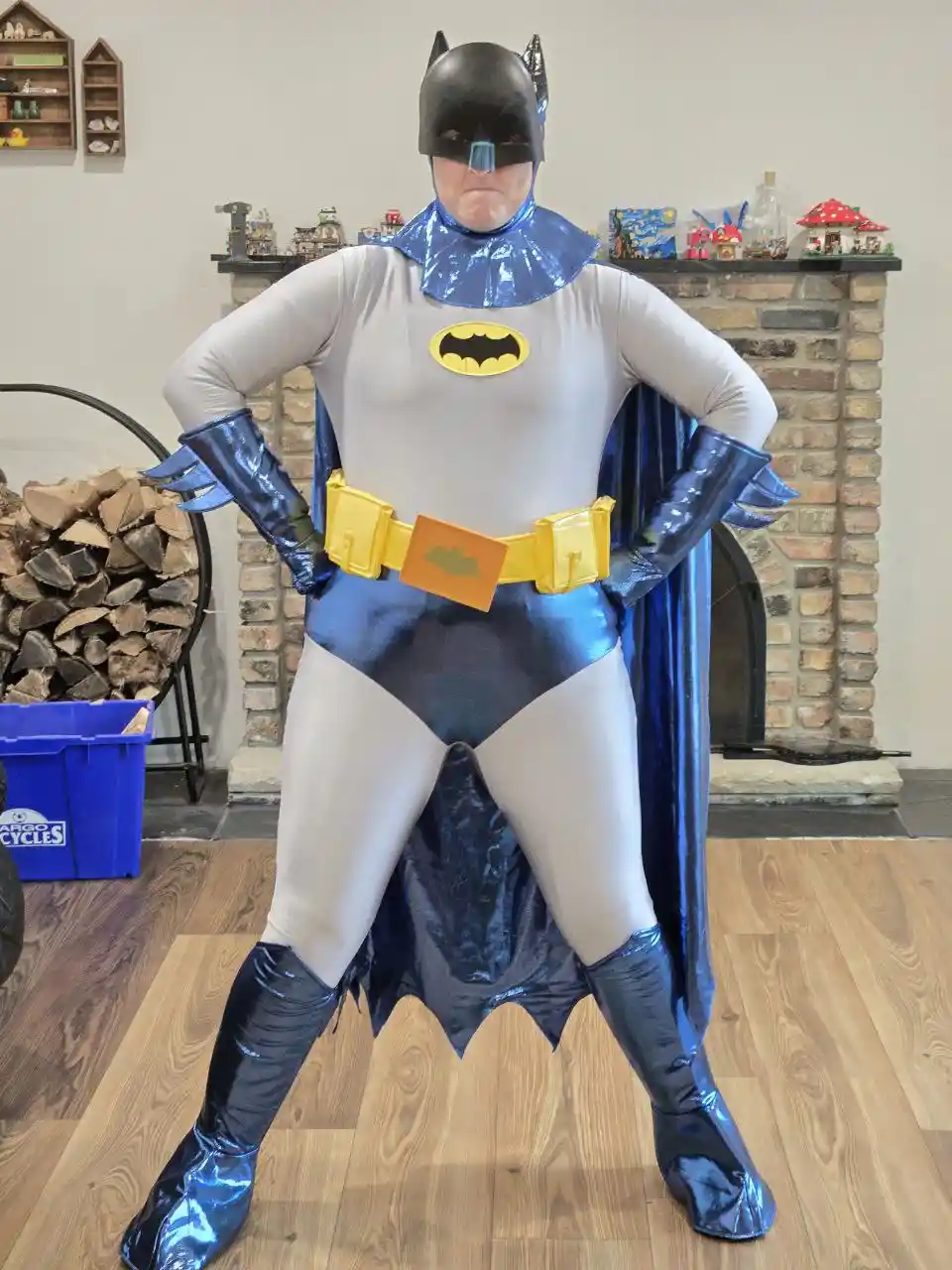 A person in a full Batman costume stands with hands on hips in front of a brick fireplace. The costume is mostly grey and…