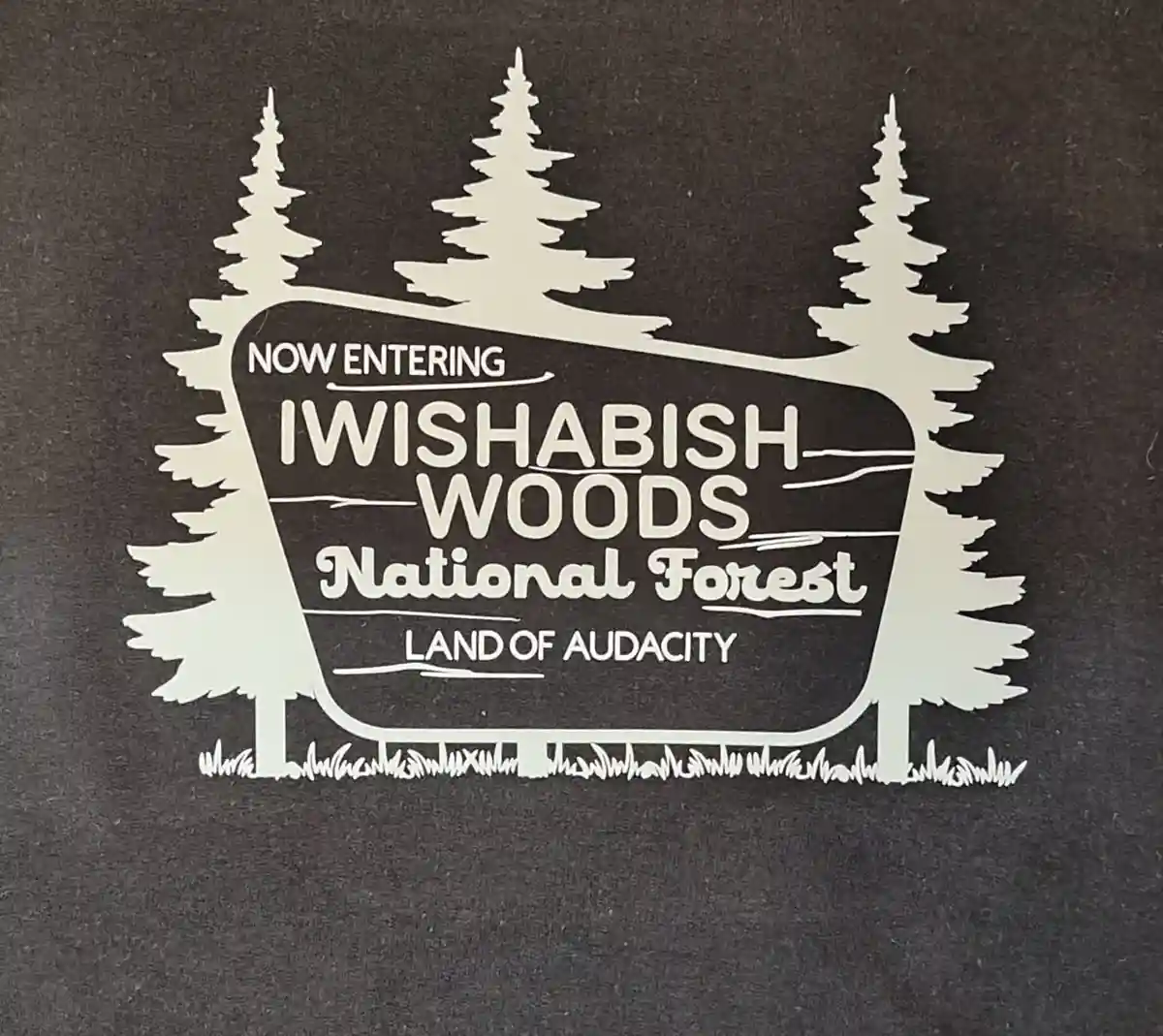 A white cutout sign on dark fabric depicts a forest entrance with three pine trees framing a curved sign. The sign reads…