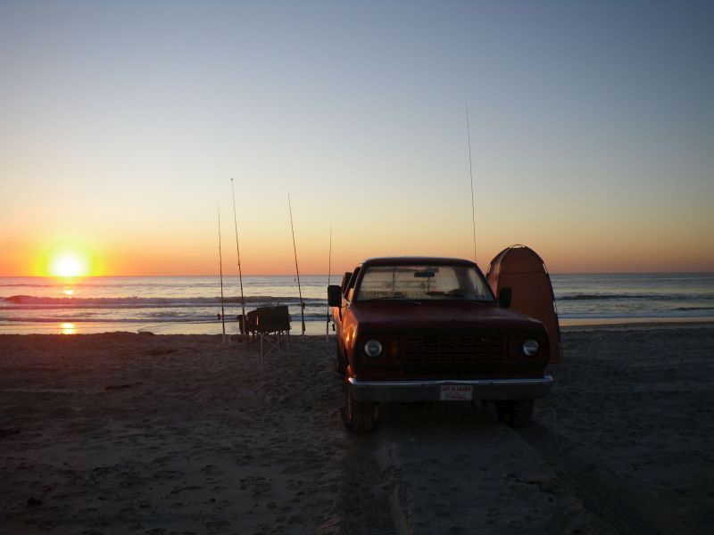 A red pickup truck is parked on a beach at sunset, with fishing rods set up nearby and a surfboard leaning against the…