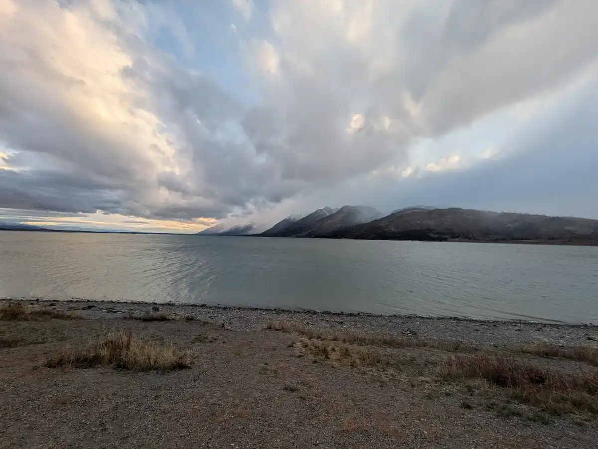A scenic photo of a calm lake with distant snow-capped mountains under a dramatic cloudy sky, with a gravelly shoreline in…