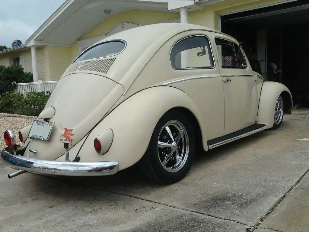 A cream-colored classic Volkswagen Beetle is parked on a concrete driveway in front of a house with an open garage. The car…