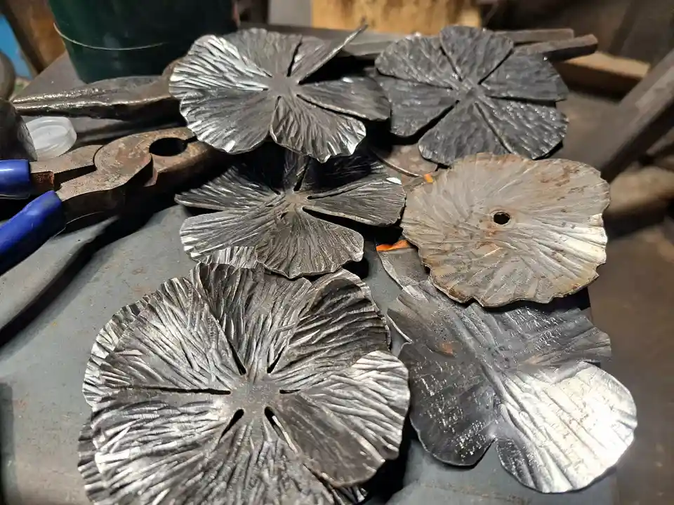 Several hand-hammered, metallic flower-shaped metal pieces with textured petals are arranged on a workbench, surrounded by…