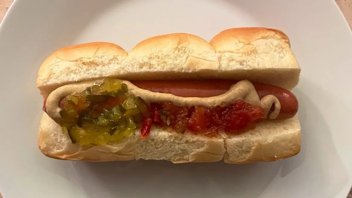A hot dog in a split bun sits on a white plate, topped with mustard, green pickles, and red salsa. The overlaid text reads…