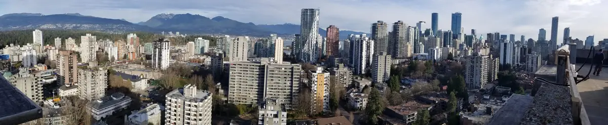 A panoramic photo of a dense urban skyline with numerous high-rise buildings, distant snow-capped mountains, and a partly…