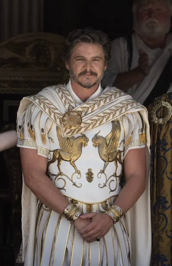 A man in ornate white and gold ancient Greek-style armor with griffin motifs stands with hands clasped, looking unimpressed…