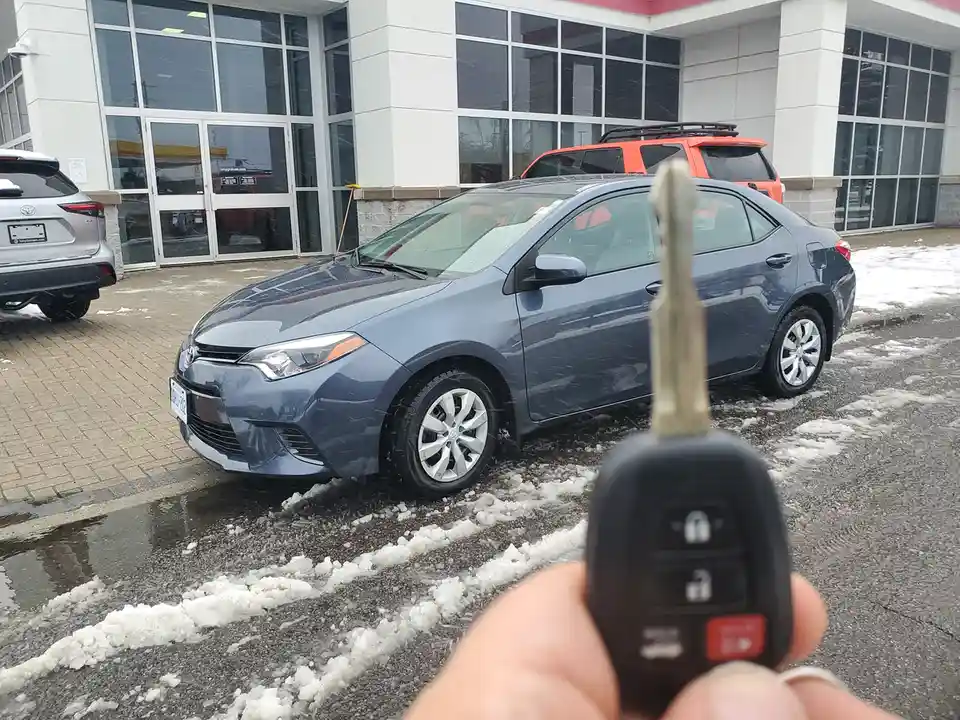 A first-person perspective shows a hand holding a car key fob in the foreground, with a blue Toyota Corolla parked outside a…