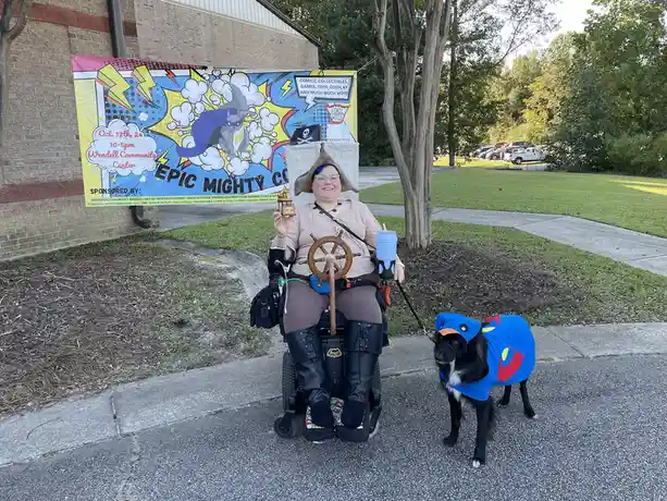 A person in a wheelchair, wearing a pirate hat and holding a ship's wheel and a small trophy, poses with their dog dressed…
