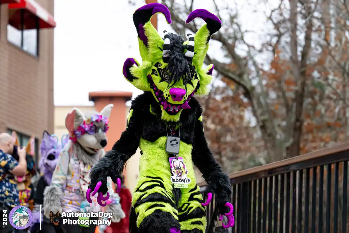 A person in a vibrant green and black fursuit with purple horns and claws stands prominently, wearing a name tag that reads…