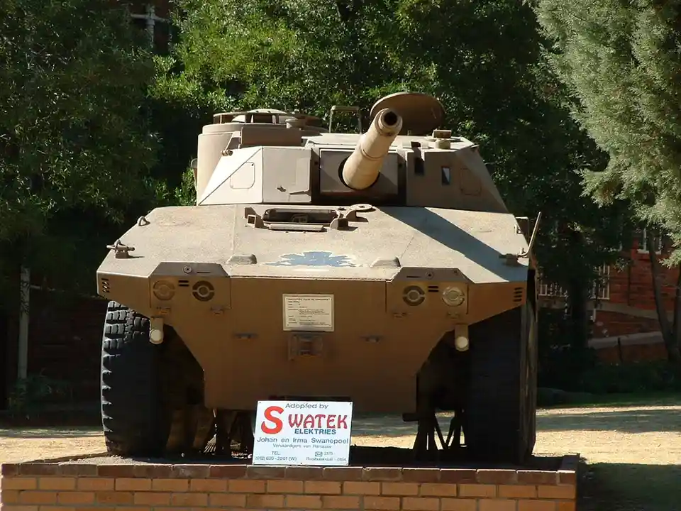 A tan armored vehicle, likely a military tank or armored personnel carrier, is displayed outdoors on a brick pedestal…
