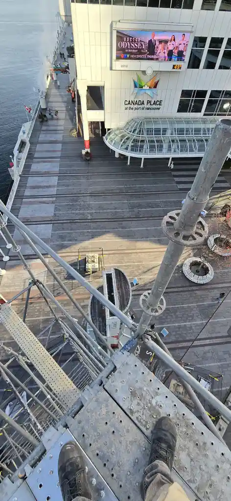 A first-person view from atop a tall metal scaffolding platform, showing a worker's feet and legs at the edge. Below is a…