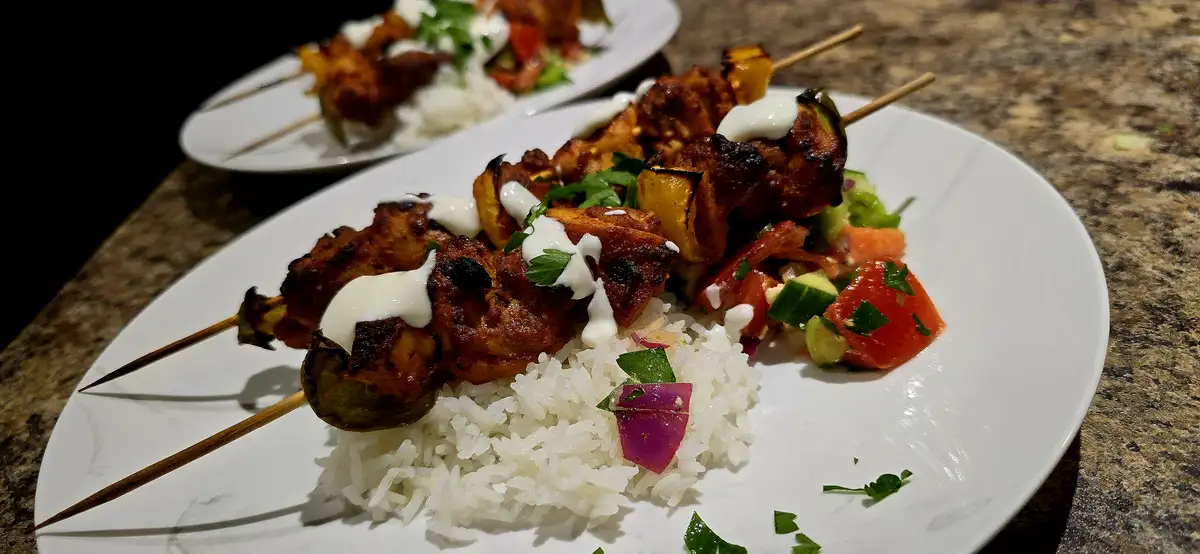 A close-up photo of two plates of grilled chicken and vegetable skewers served over white rice with a side of chopped…