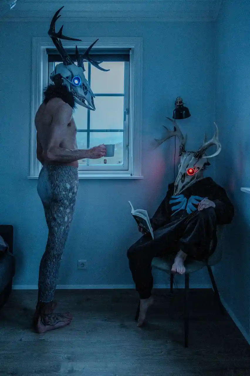 Two people wearing deer skull masks with glowing eyes stand in a dimly lit room; one stands by a window holding a mug, the…