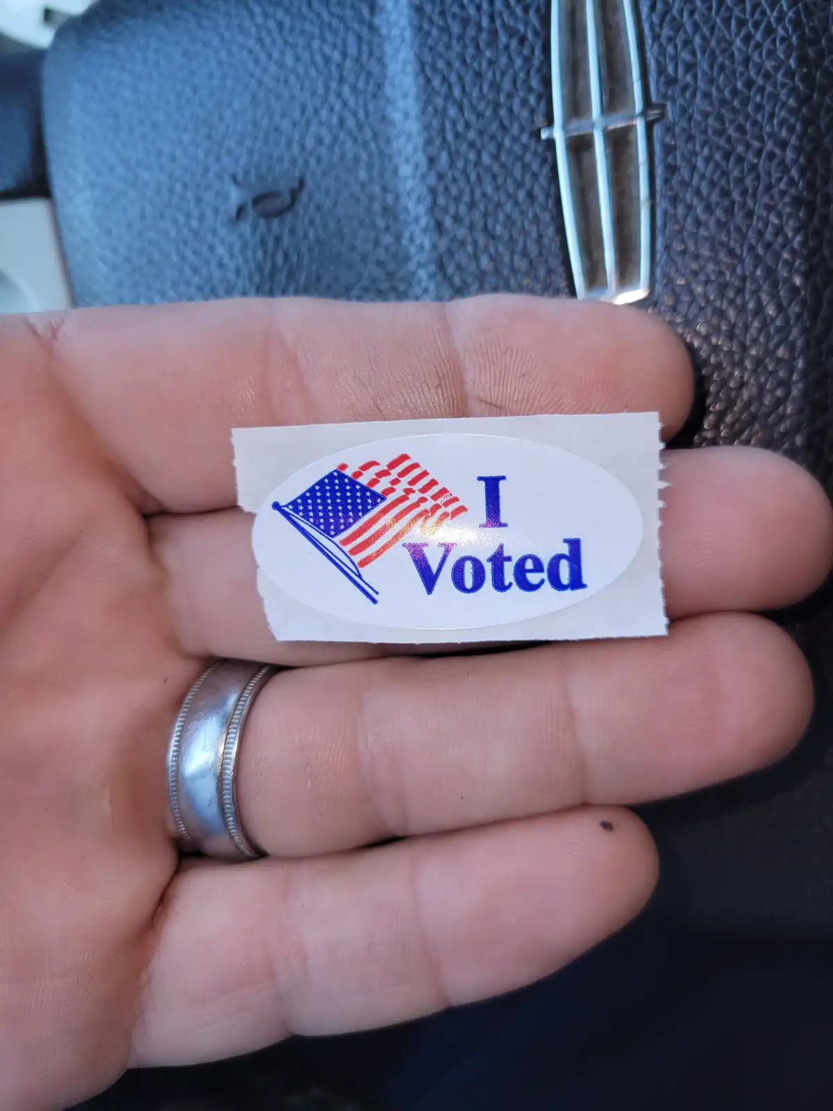 A close-up photo shows a person's hand holding a small oval sticker with the text 'I Voted' in blue, next to a stylized…