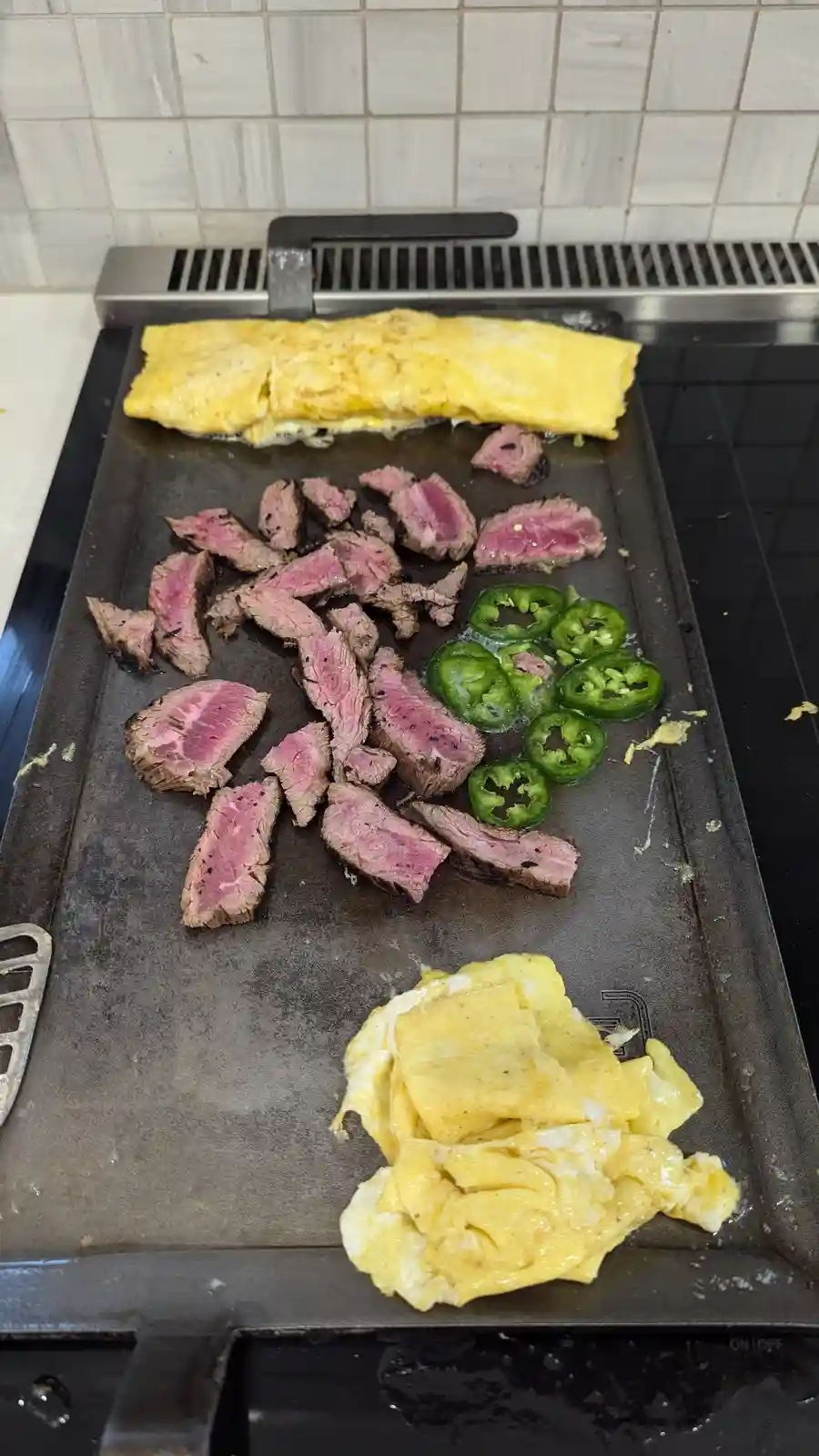 A griddle shows cooked steak slices, sliced jalapeños, and two forms of scrambled eggs—one folded and one loose—on a…