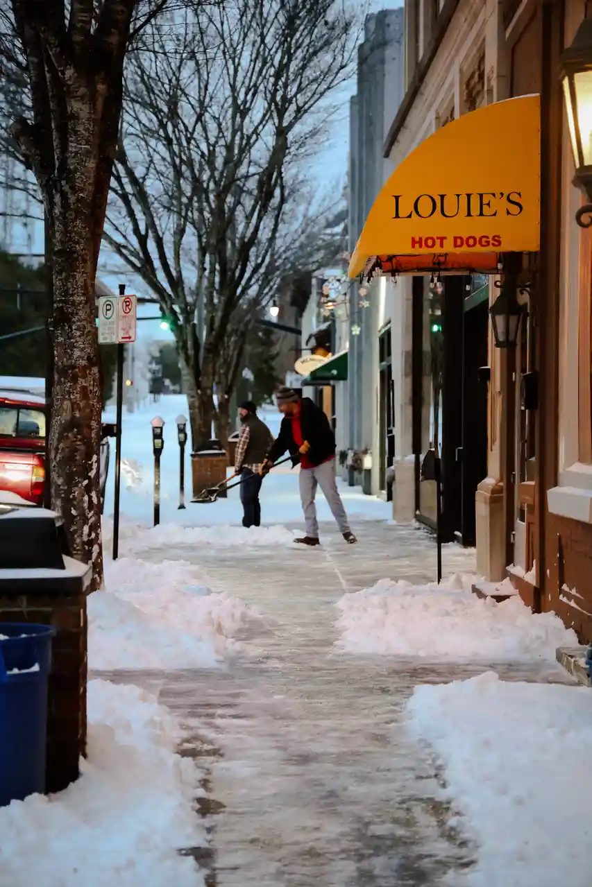 Two men shovel snow off a sidewalk in front of a storefront with a yellow awning that reads 'LOUIE'S HOT DOGS'. The sidewalk…