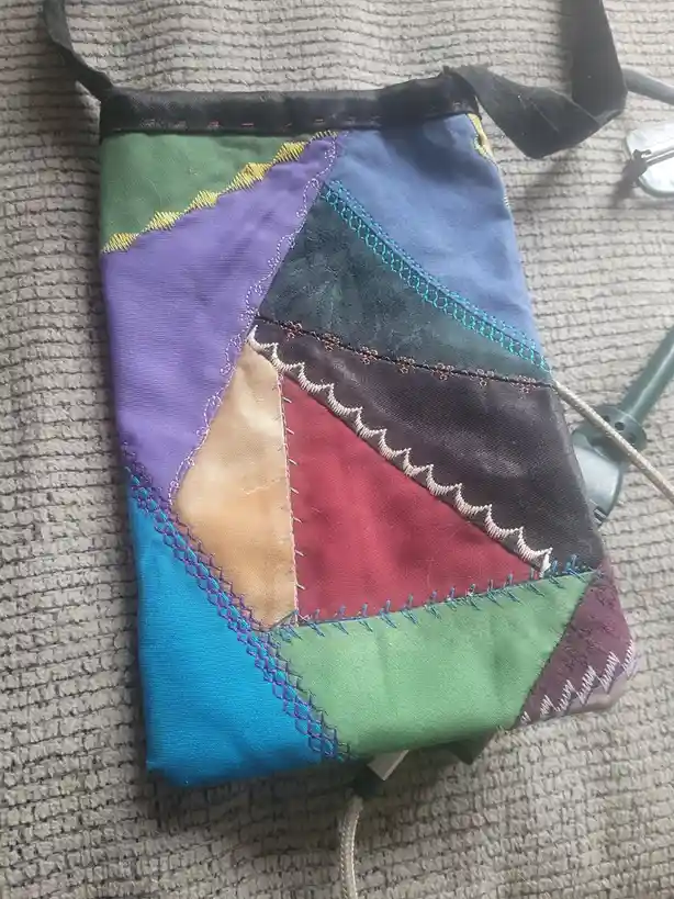 A handmade patchwork bag made from various colorful fabric scraps, stitched together with different decorative seams, rests…