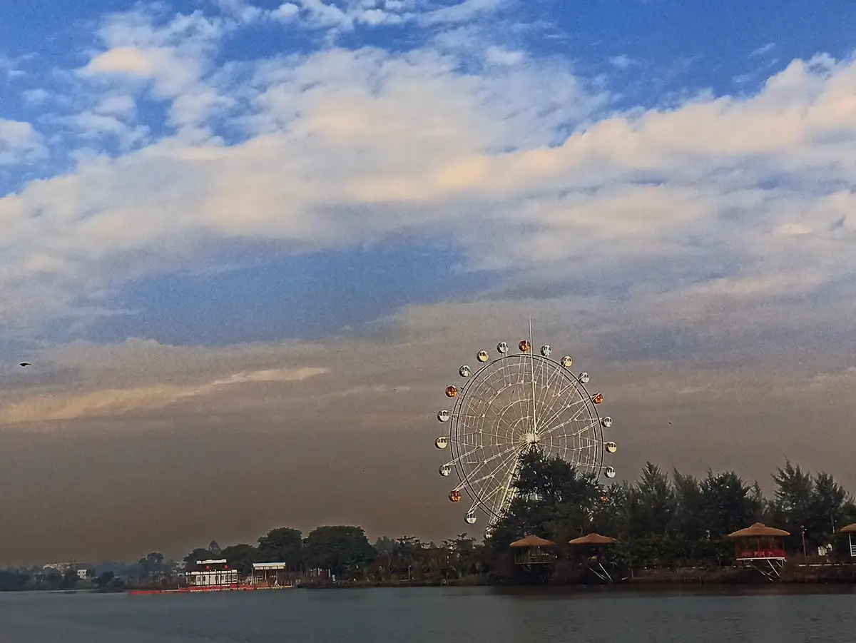 A scenic photo of a large white Ferris wheel by a lake under a partly cloudy sky, with trees and small pavilions along the…