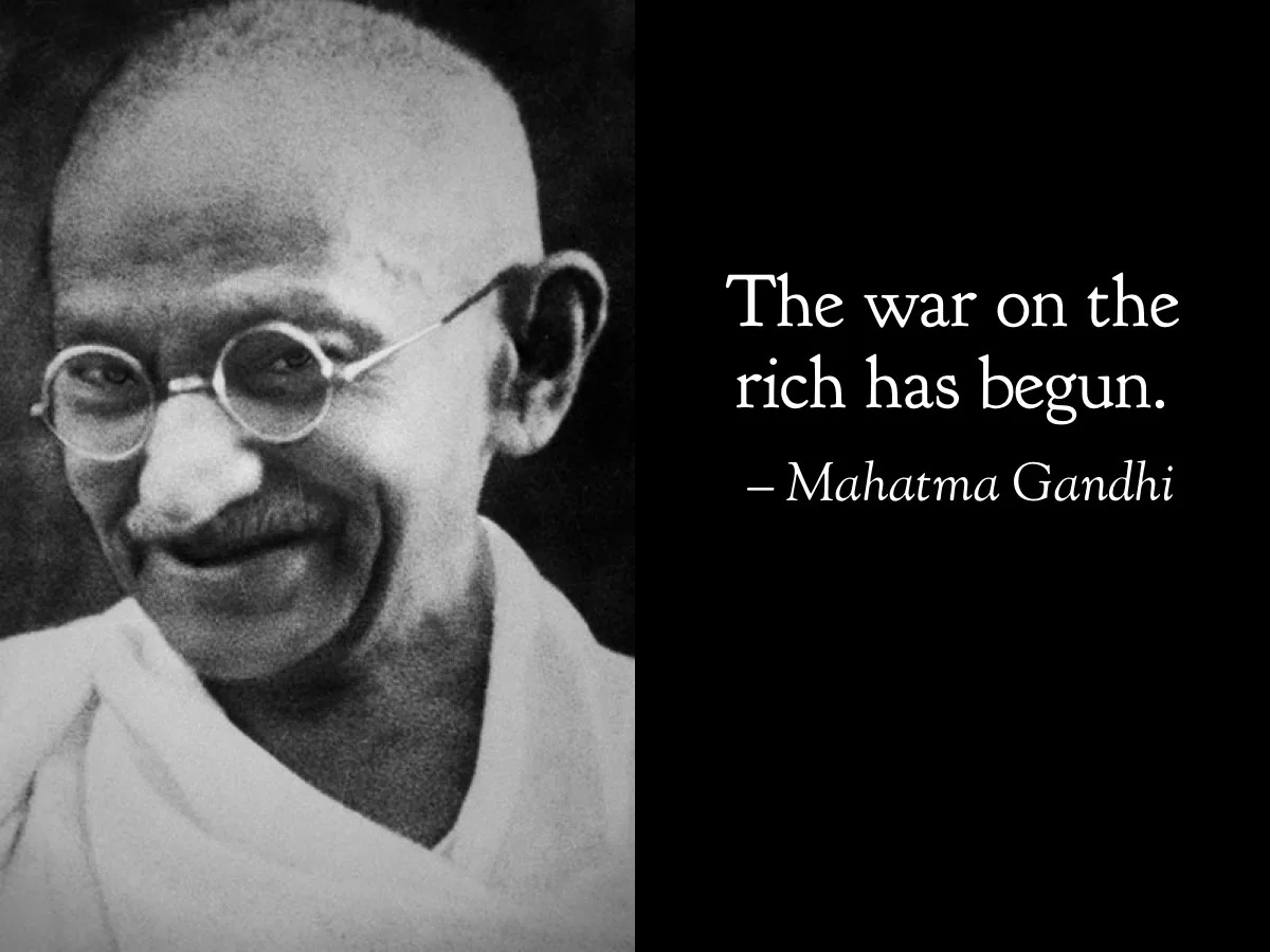 A black-and-white photo of Mahatma Gandhi smiling, wearing round glasses and a white garment, with white text on a black…