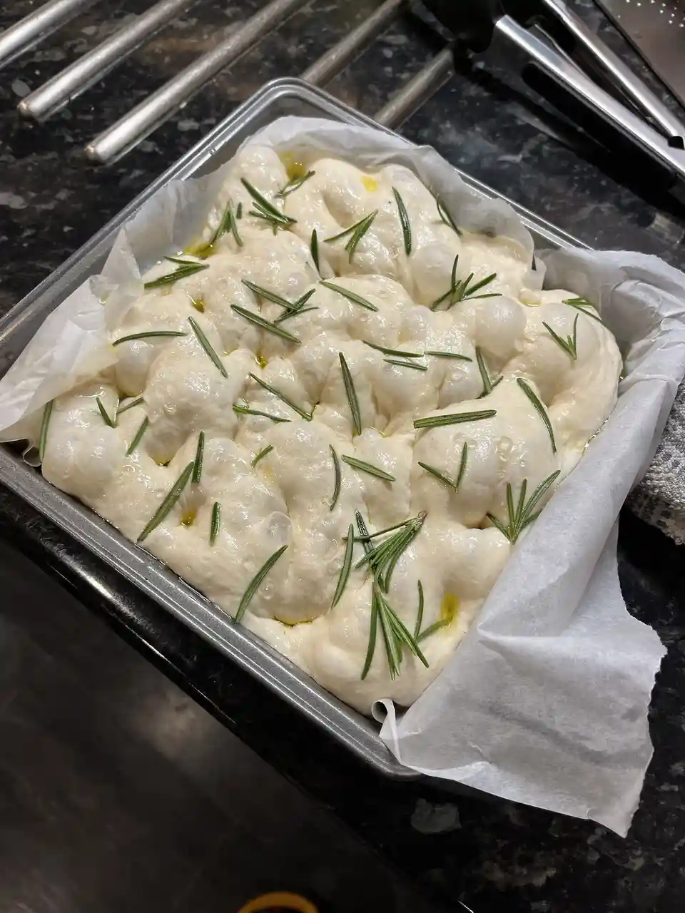 A close-up photo shows a rectangular metal baking pan lined with parchment paper, containing risen, bubbly white dough…