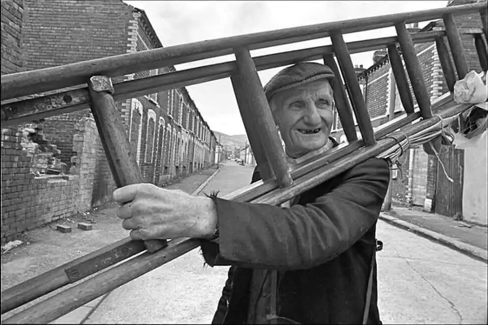 A black-and-white photo shows an elderly man with a wide, toothy grin, carrying a long wooden ladder over his shoulder down…