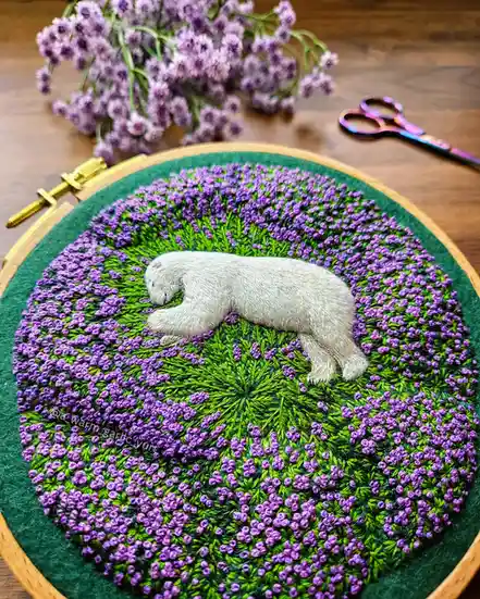 A detailed embroidery in a wooden hoop shows a small, gray, sleeping polar bear lying on a bed of green grass and purple…