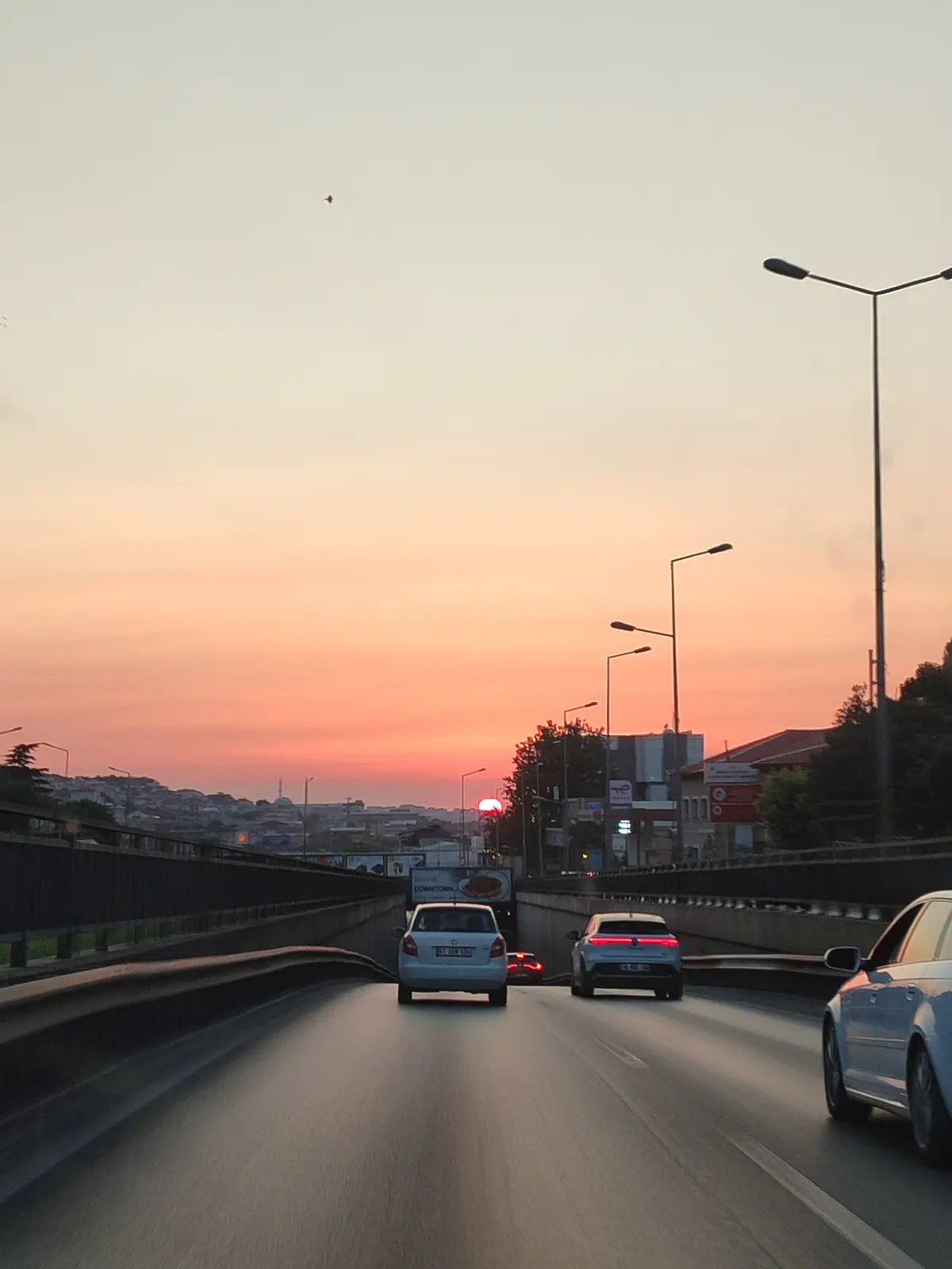 A photo taken from a car on a highway at sunset, showing several cars ahead, streetlights, and a cityscape in the distance…