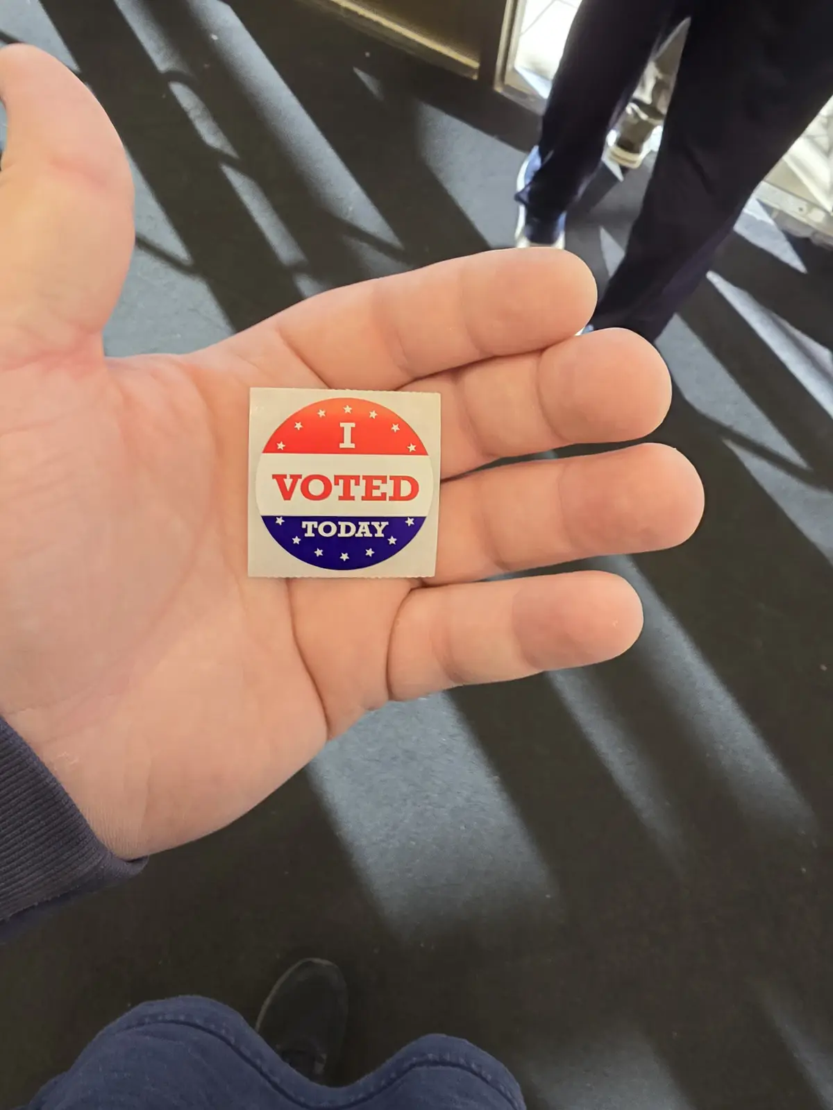 A person holds up a small, square 'I VOTED TODAY' sticker in their open palm. The sticker features red, white, and blue…
