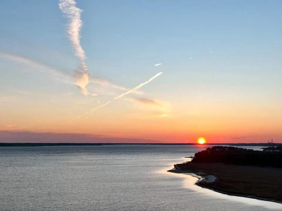 A serene sunset over water with a distant shoreline, featuring a long contrail in the sky that resembles a tear falling from…