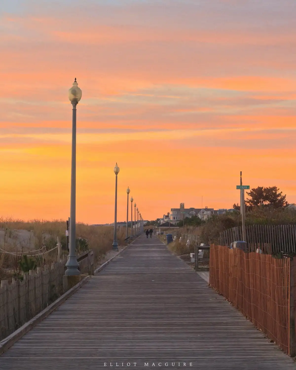 A serene wooden boardwalk stretches into the distance under a vibrant orange and pink sunset sky, lined with lampposts and…