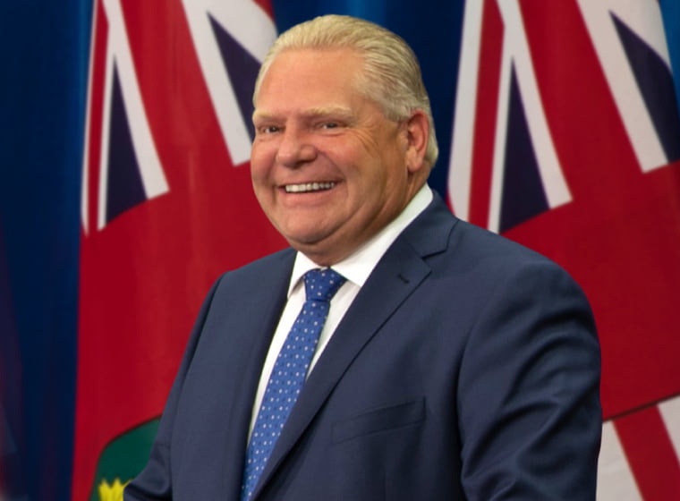 a man in a blue suit and blue polka-dot tie smiles broadly in front of multiple red, white, and blue flags. The overlaid…