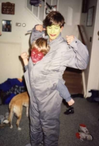 Two children in a home setting, one wearing a green face paint and a grey jumpsuit, holding up the other child who has red…