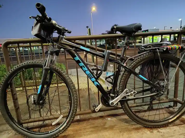 A black Trek mountain bike with blue accents is parked against a metal railing at dusk, equipped with a water bottle…