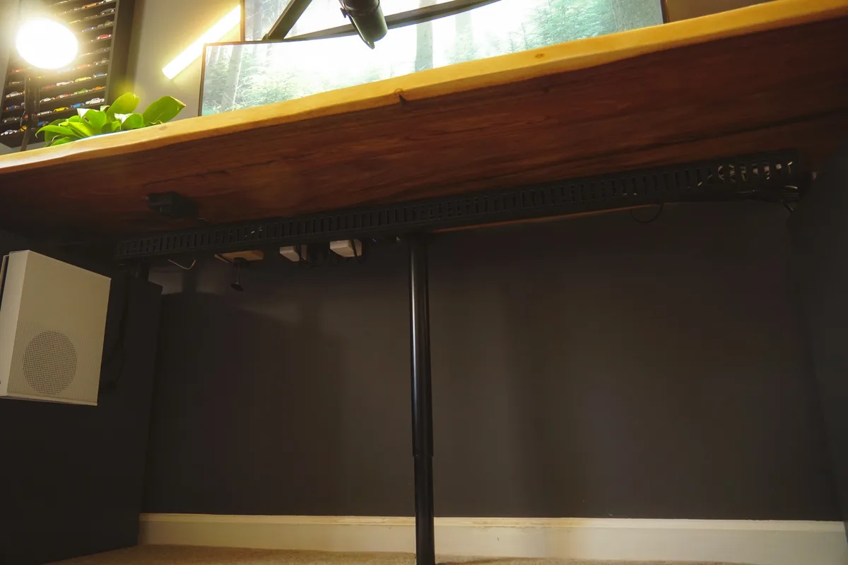 A low-angle photo shows the underside of a thick, rustic wooden desk supported by a single black metal pole. Below the desk…