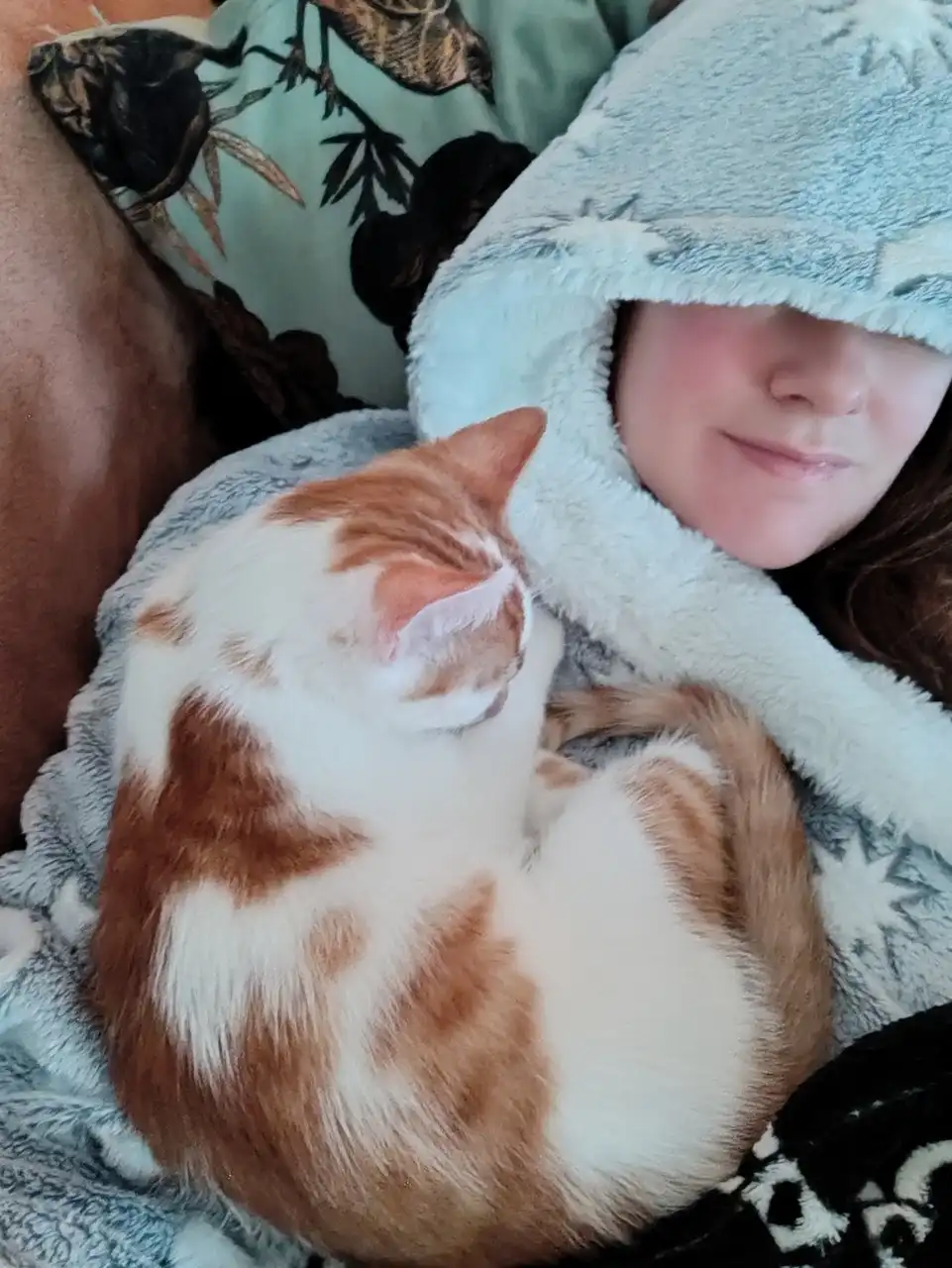 A person wearing a light blue, fluffy hooded blanket smiles contentedly while cuddling a curled-up orange and white cat. The…
