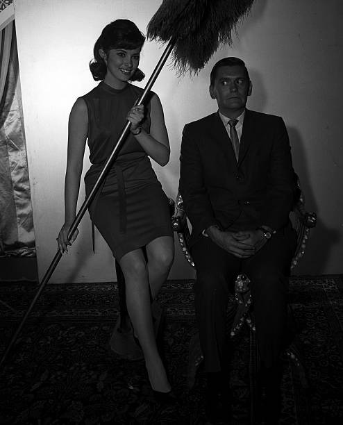 A black-and-white photo shows a smiling woman in a 1950s-style dress holding a large feather duster while standing next to a…