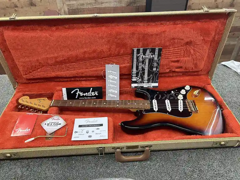 A Fender Stratocaster electric guitar with a sunburst finish sits inside its open, red-furred hardshell case. The case…
