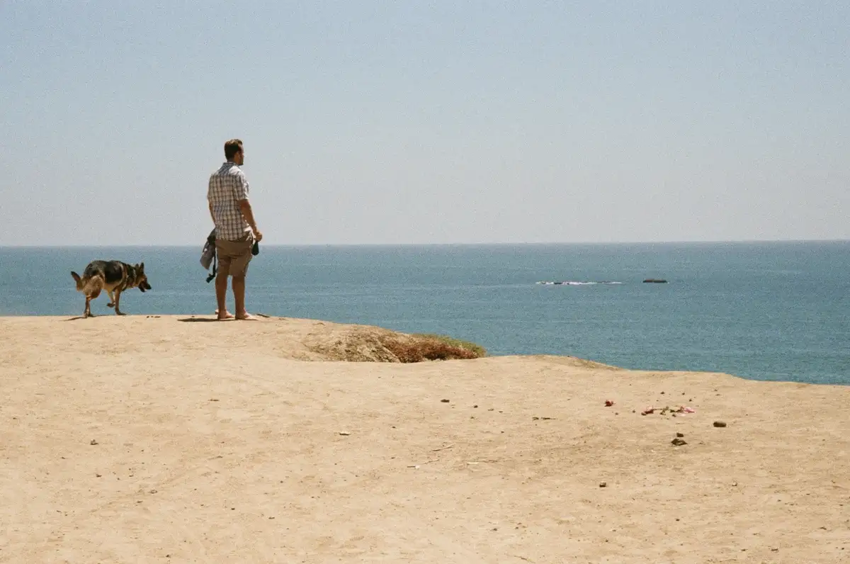 A man in a plaid shirt and shorts stands barefoot on a sandy cliff overlooking the ocean, with a German Shepherd dog beside…